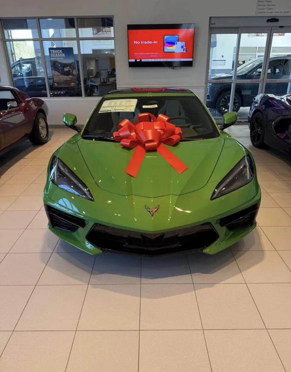 A green Chevrolet Corvette C8 with a large red bow on the hood, displayed in a car dealership showroom.