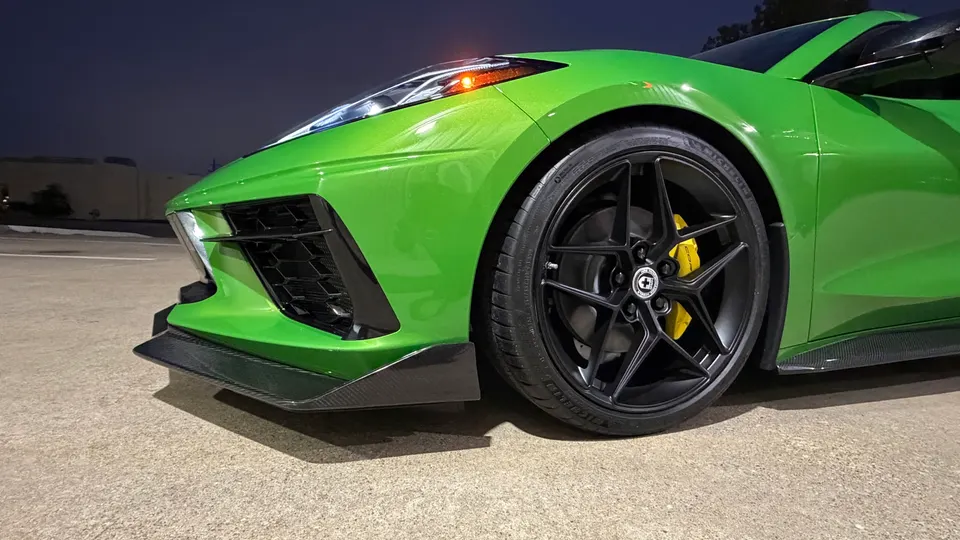 Close-up of a bright green C8 Corvette's front corner, showcasing custom black wheels, yellow brake calipers, and a carbon fiber front splitter at night.