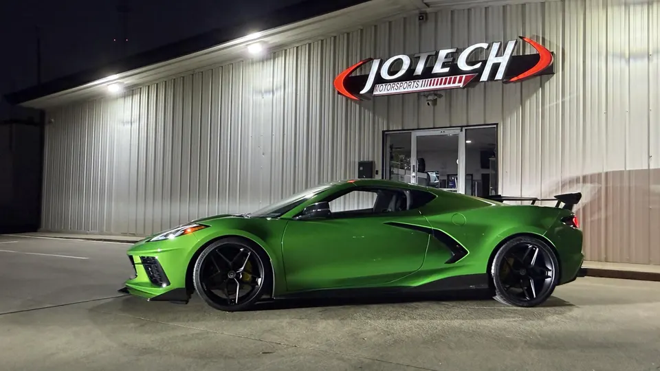 Green Chevrolet Corvette C8 with aftermarket wheels and rear wing parked outside Jotech Motorsports shop at night.