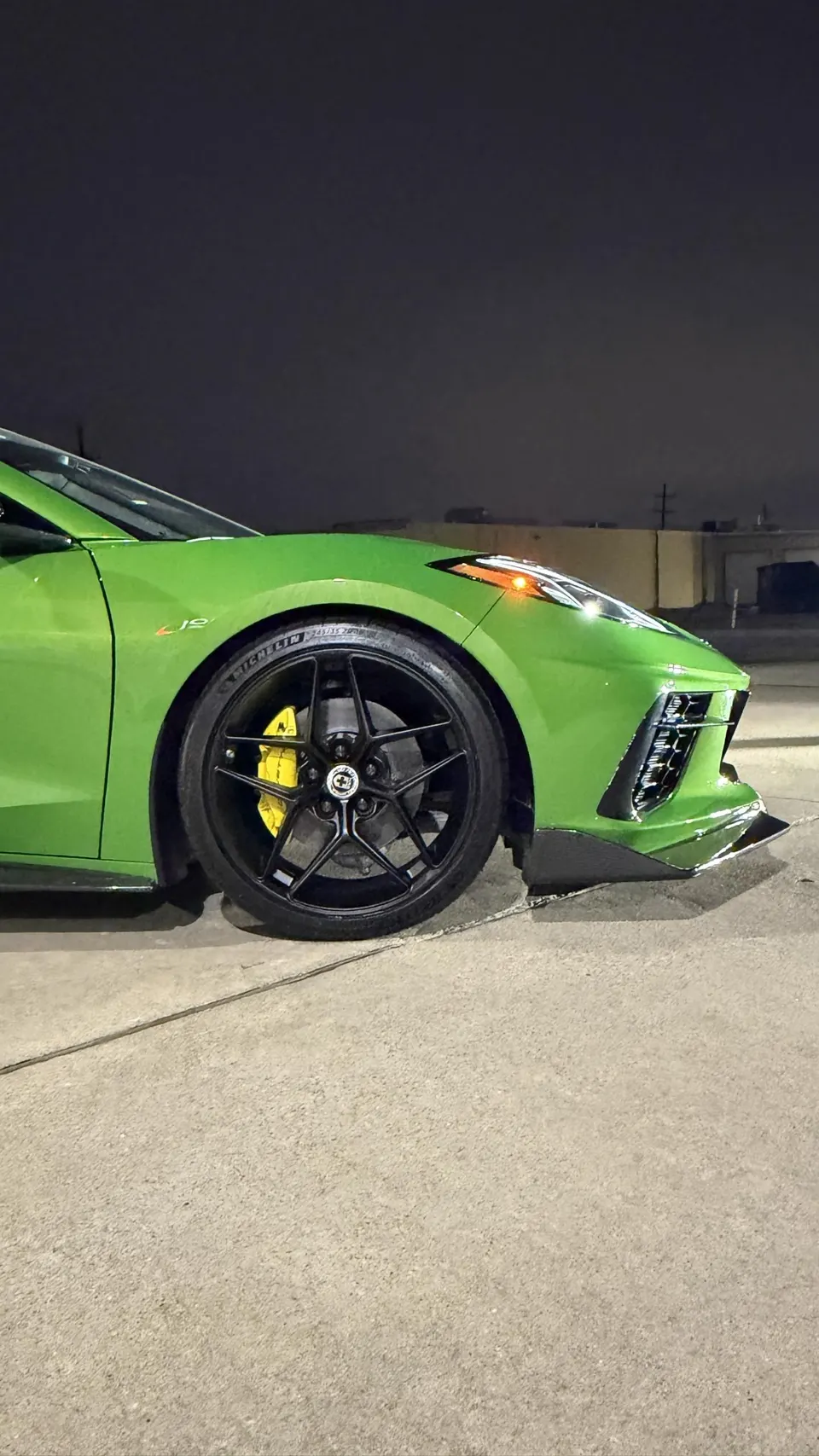 A close-up of a green C8 Corvette's front wheel, featuring black HRE wheels, yellow brake calipers, and a carbon fiber splitter at night.