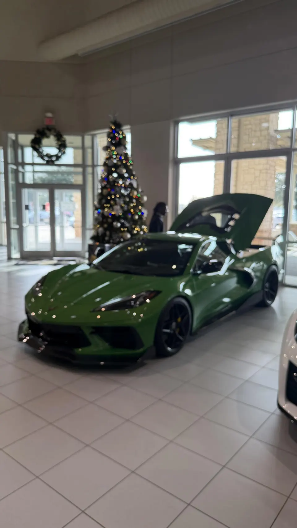 A green Chevrolet Corvette C8 with its engine cover open, displayed in a dealership showroom with a Christmas tree.