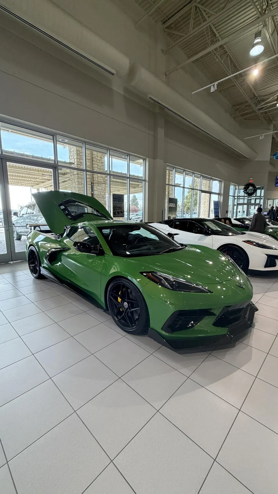 A vibrant green Chevrolet Corvette C8 Z06 with its engine cover open, displayed prominently in a bright dealership showroom.