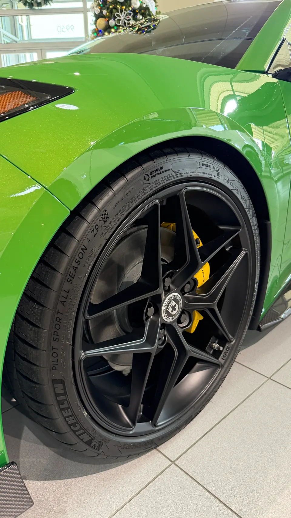 Close-up of a bright green car's front wheel, featuring black HRE wheels, Michelin tires, and yellow brake calipers in a showroom.