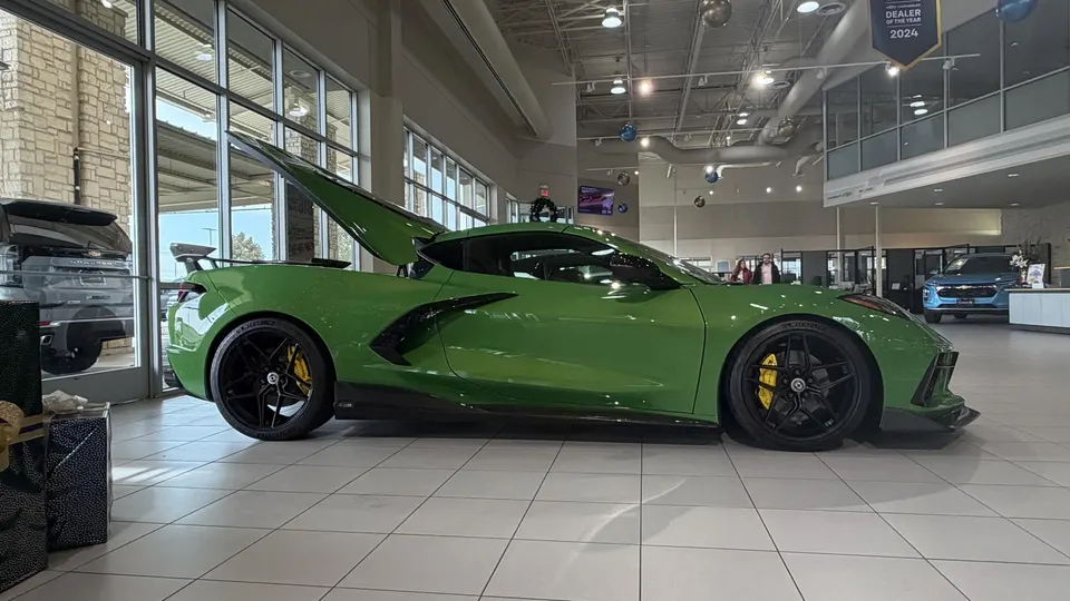 A bright green Chevrolet Corvette C8 with its rear engine cover open, displayed prominently in a modern dealership showroom.