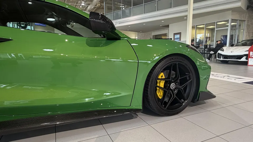Green Chevrolet Corvette C8 with black HRE wheels, yellow brake calipers, and carbon fiber side skirts in a dealership showroom.