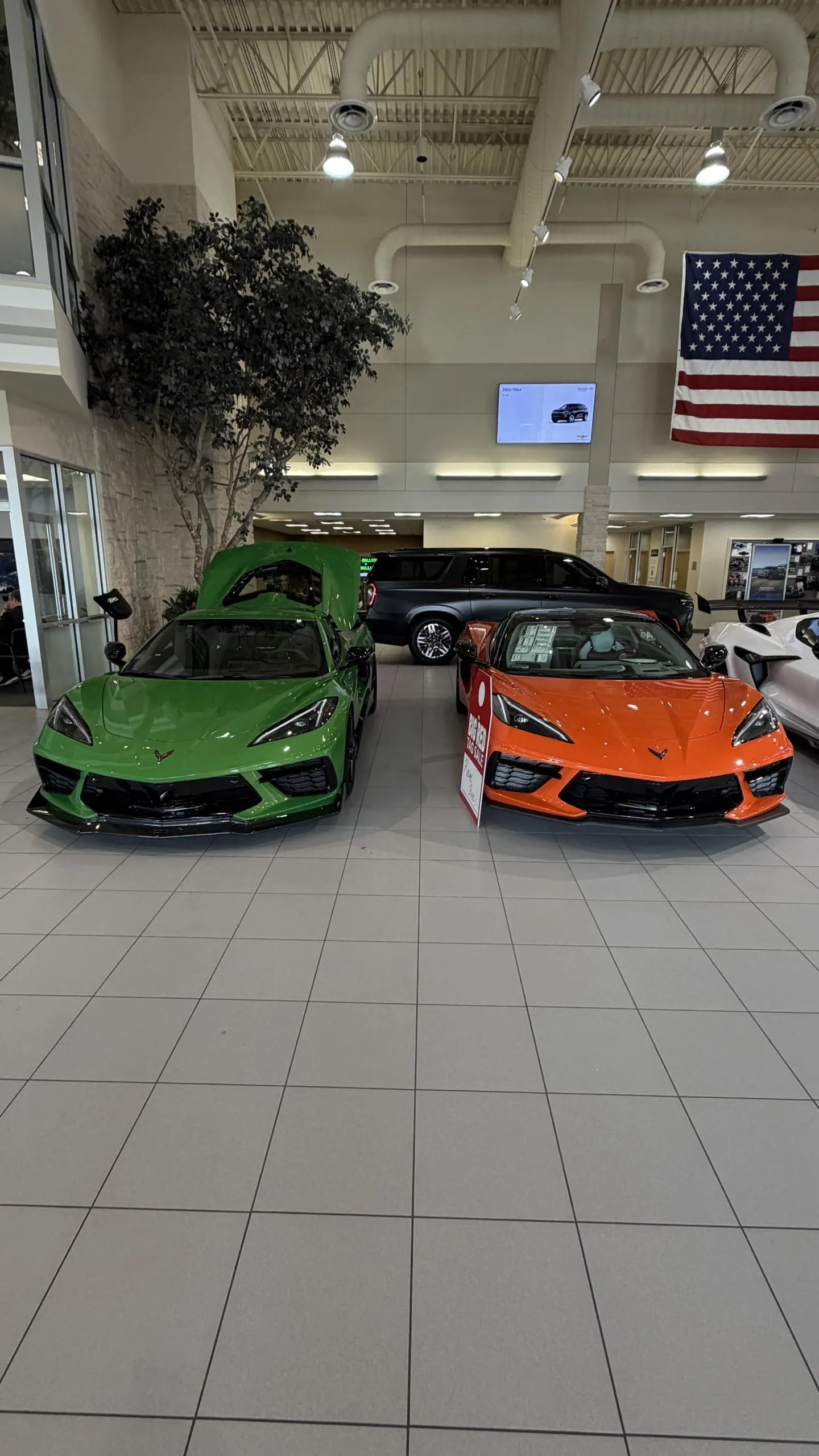 Two new Chevrolet C8 Corvettes, one green and one orange, displayed in a car dealership showroom.