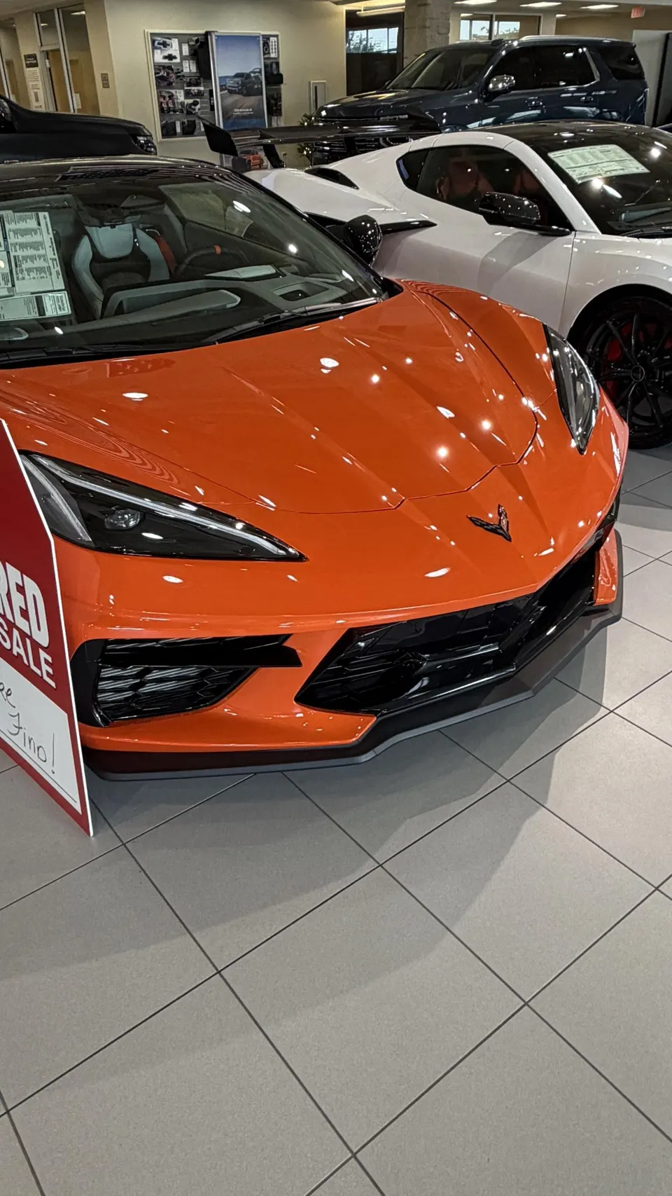 Orange Chevrolet Corvette C8 sports car on display in a dealership showroom with a 'RED SALE' sign.