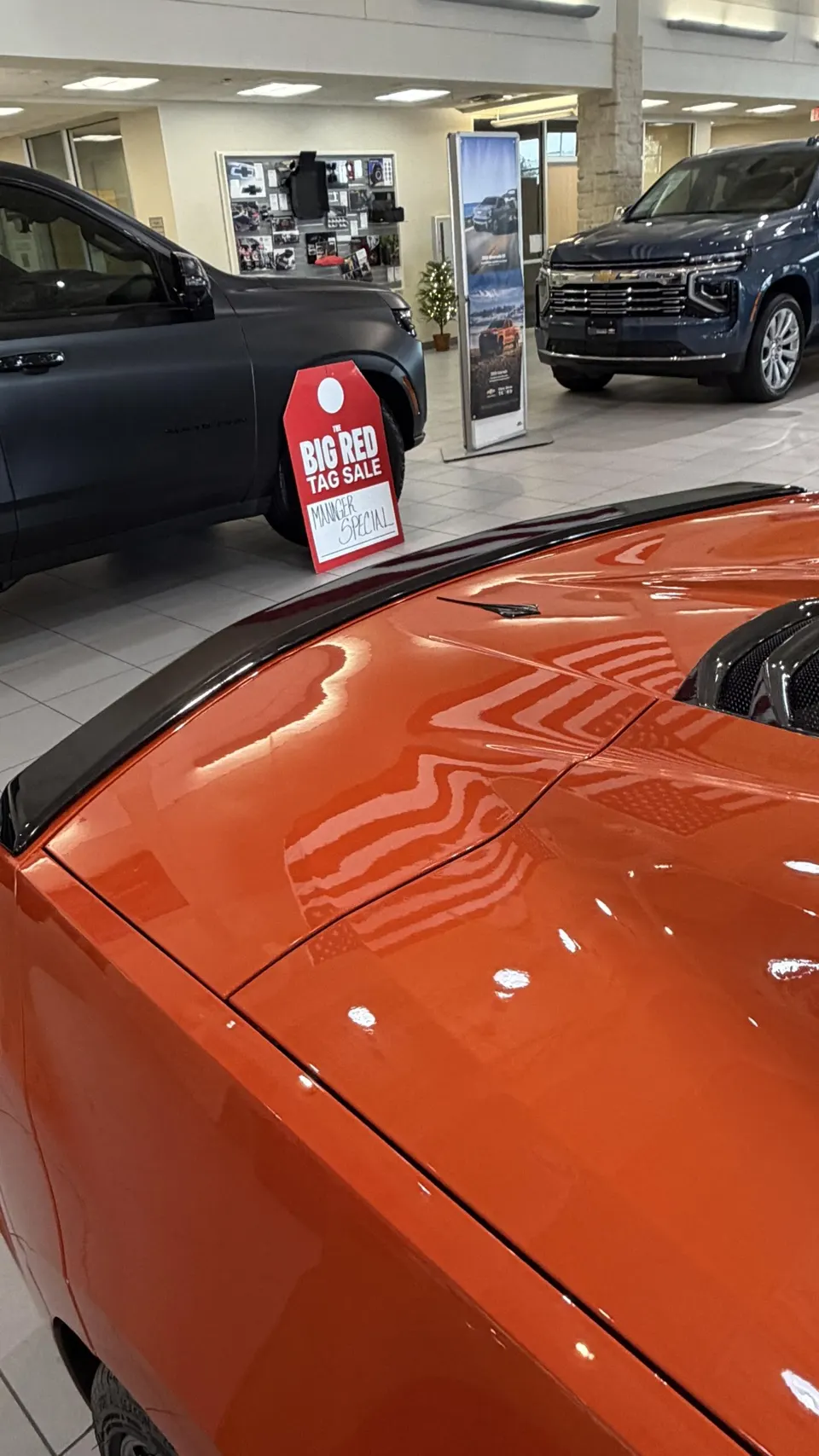 Orange Corvette C8 with a black spoiler on display in a car dealership showroom, showing reflections on its glossy paint.
