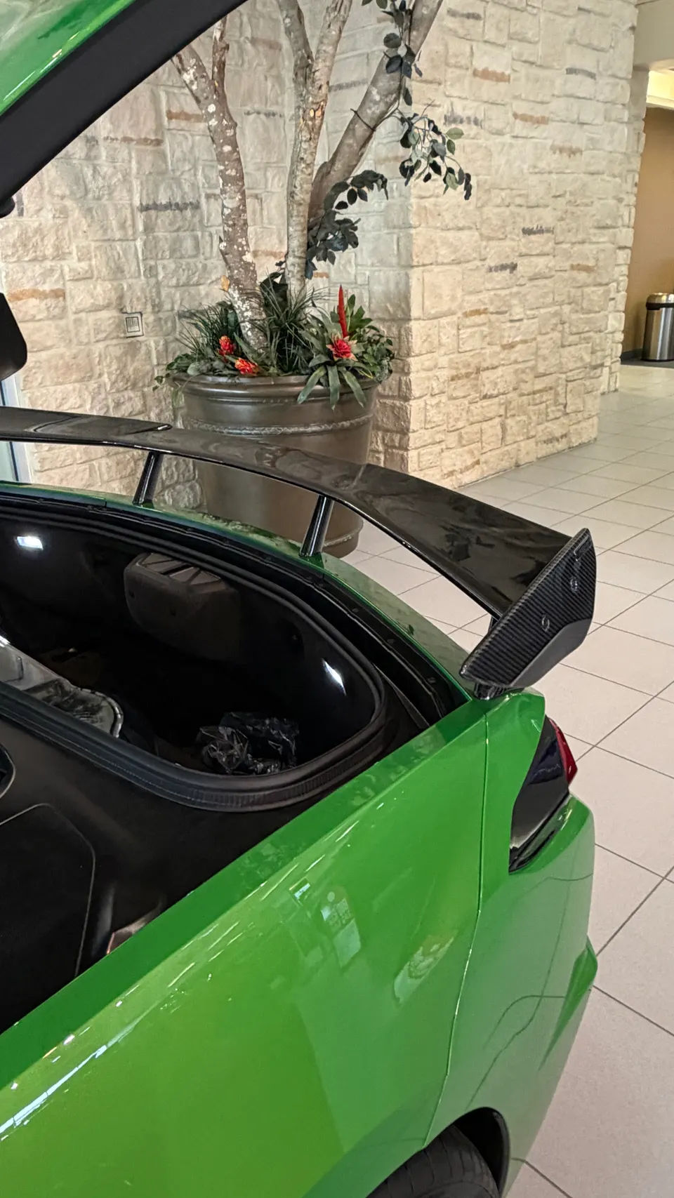 A vibrant green car with its trunk open, showcasing a prominent carbon fiber rear wing in a showroom setting.
