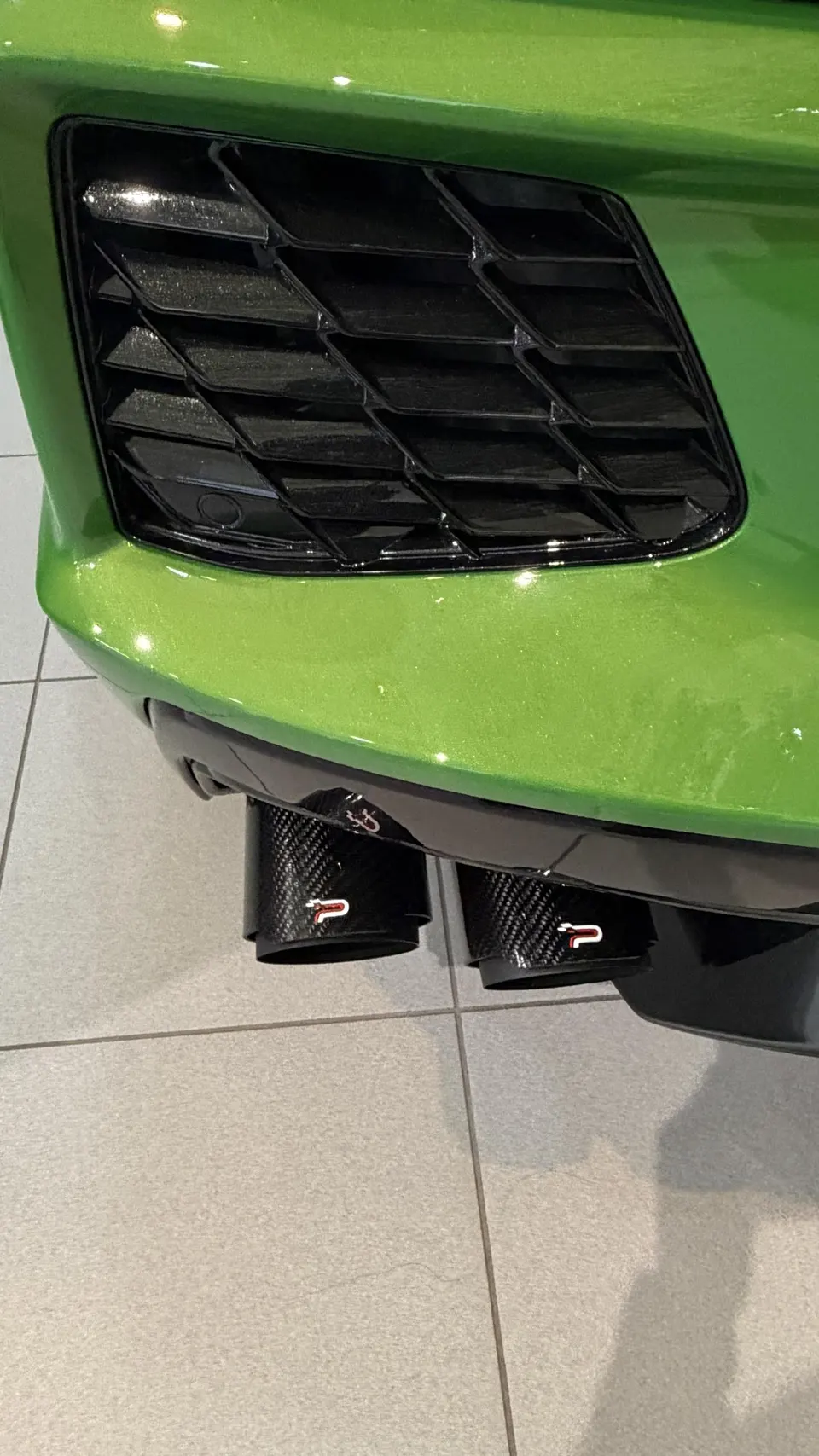 Close-up of the rear of a green Porsche, showcasing carbon fiber Akrapovic exhaust tips and a black rear grille.