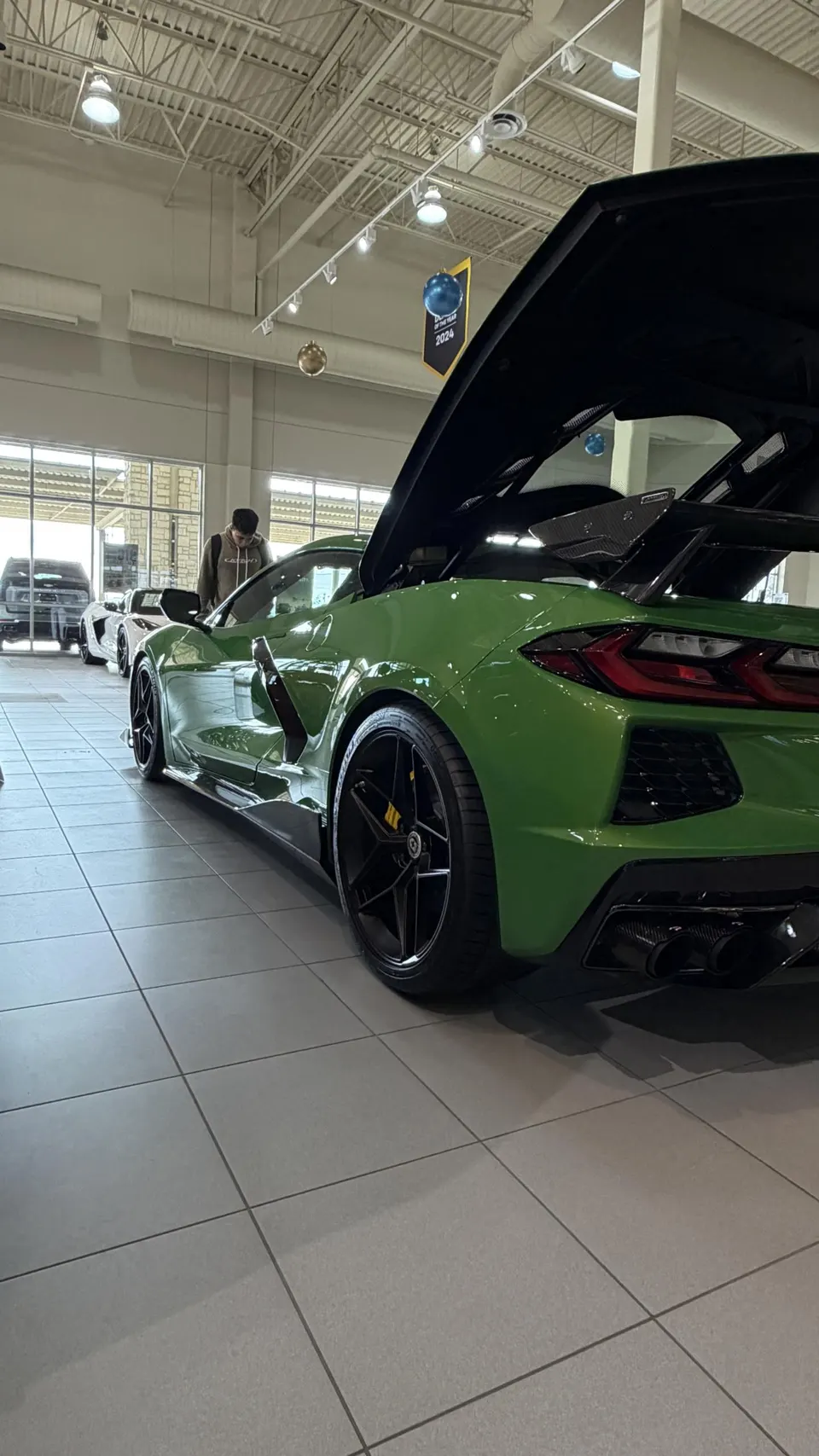 A green Chevrolet Corvette C8 Z06 with an open engine bay and carbon fiber wing on display in a dealership showroom.