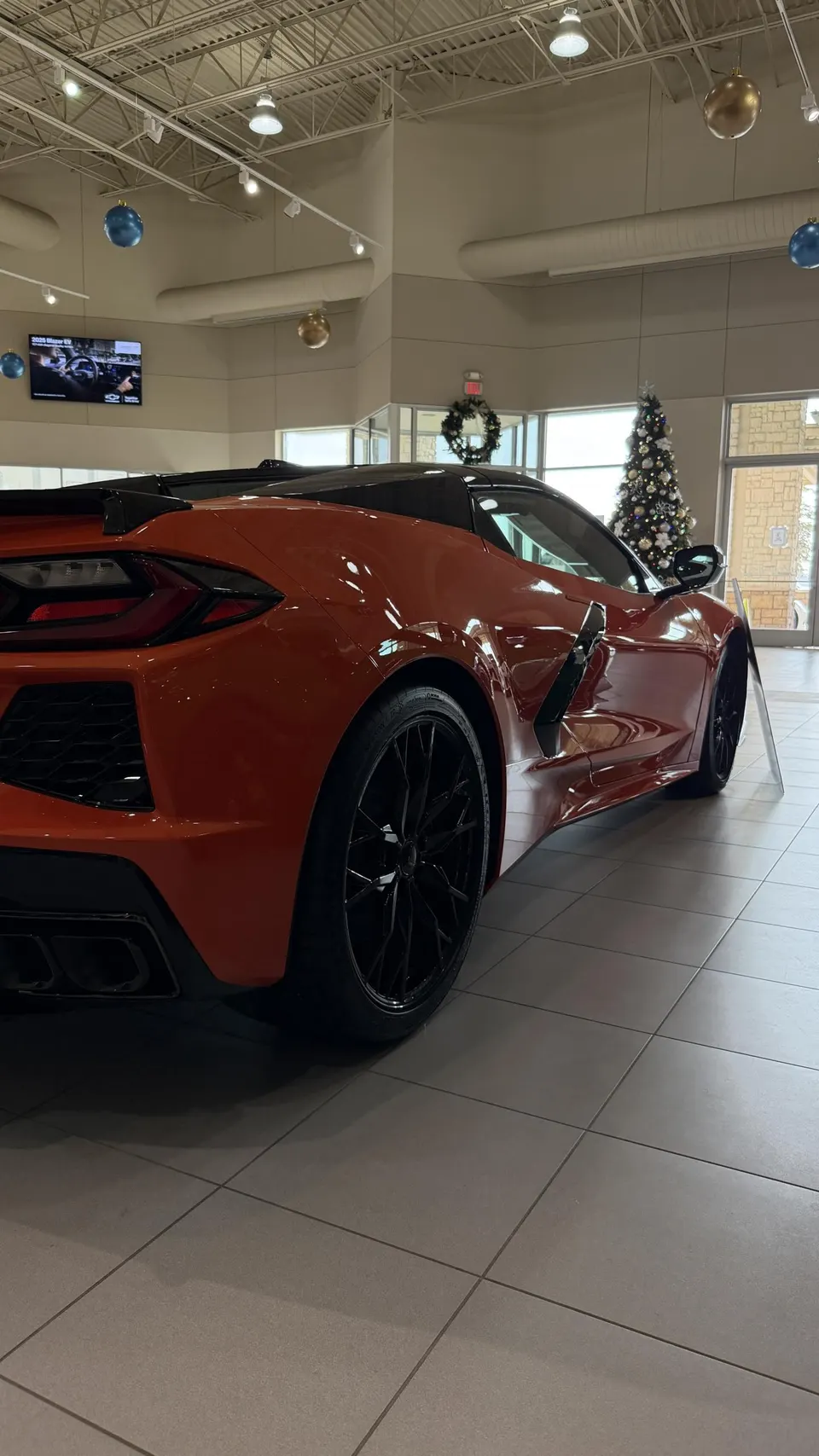 Orange Chevrolet Corvette C8 parked in a dealership showroom with Christmas decorations and tiled floor.