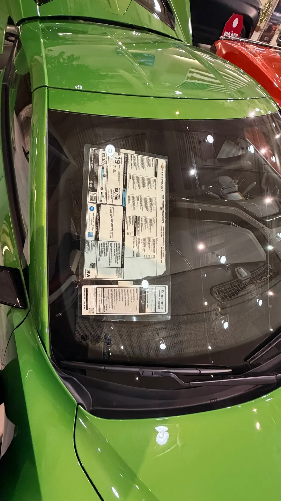 Overhead view of a green Chevrolet Corvette with a window sticker on the windshield, showcasing vehicle details in a showroom.