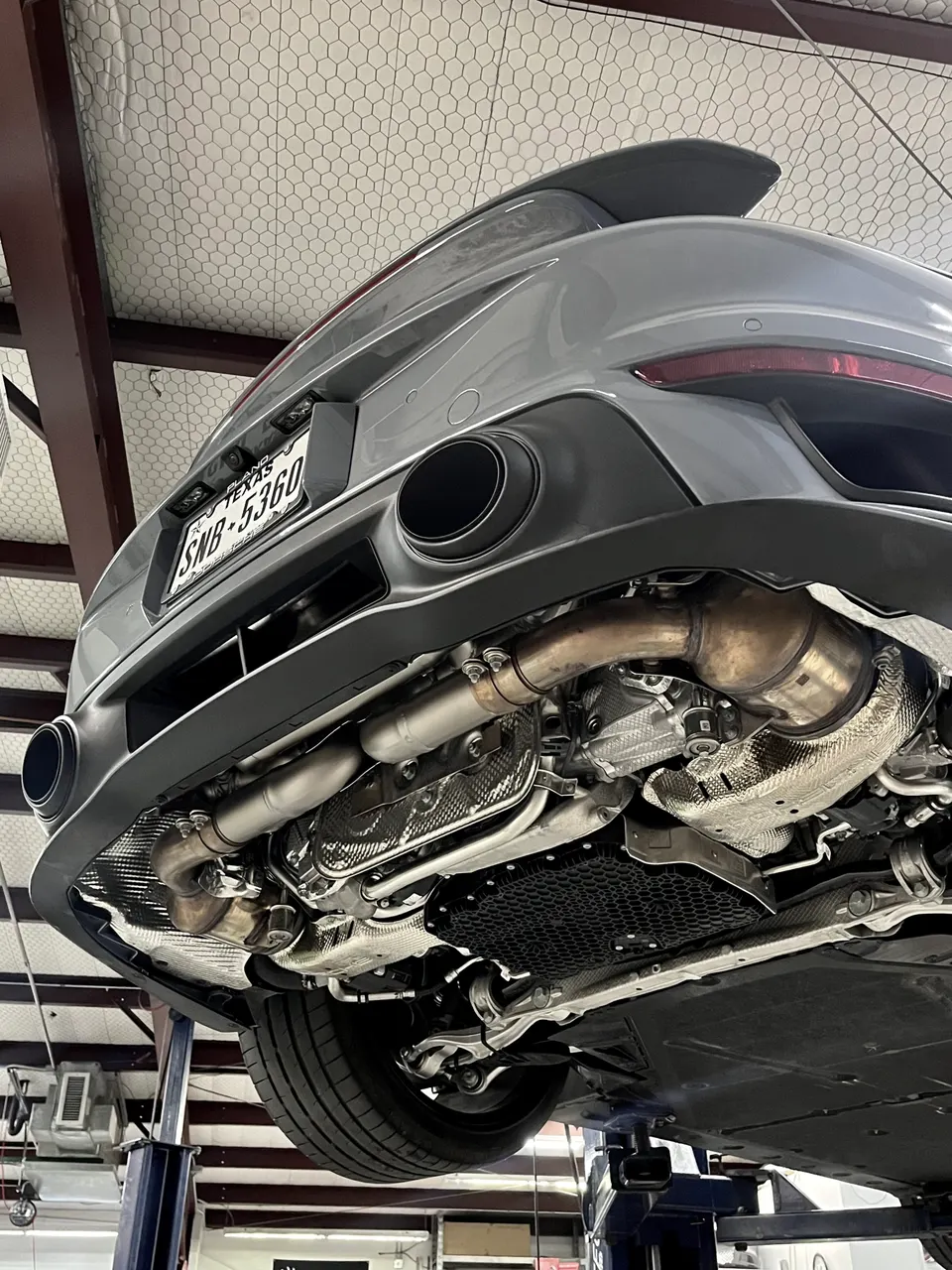 A grey Porsche sports car on a lift in a shop, showcasing its underside and exhaust system with heat shields.