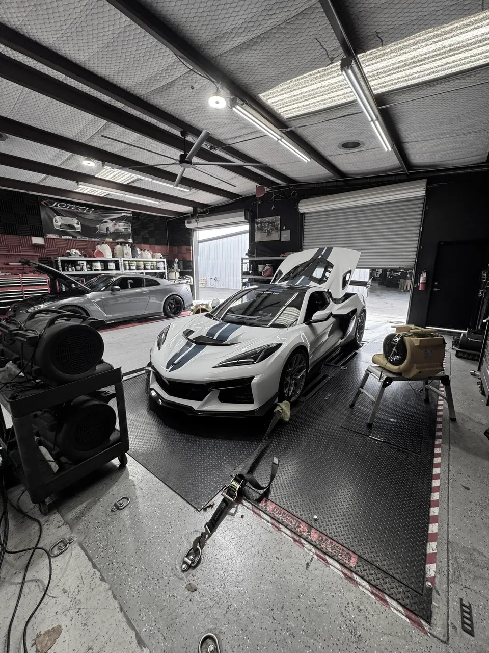 A white Corvette C8 Z06 with blue stripes strapped to a dyno at Jotech Motorsports, with a Nissan GT-R in the background.