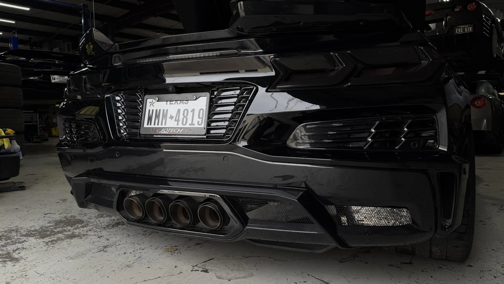 A black C8 Corvette's rear bumper and quad exhaust tips in a Jotech Motorsports shop.