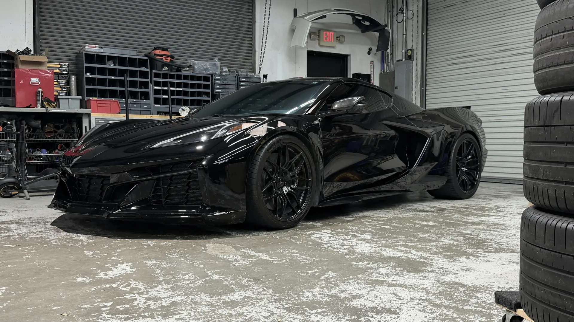 A black C8 Corvette parked in a performance tuning shop with shelves of parts and stacked tires in the background.