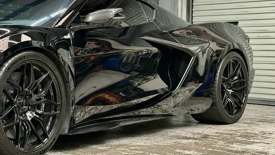 Black Chevrolet Corvette C8 with carbon fiber side skirts, mirror caps, and aftermarket forged wheels in a garage.
