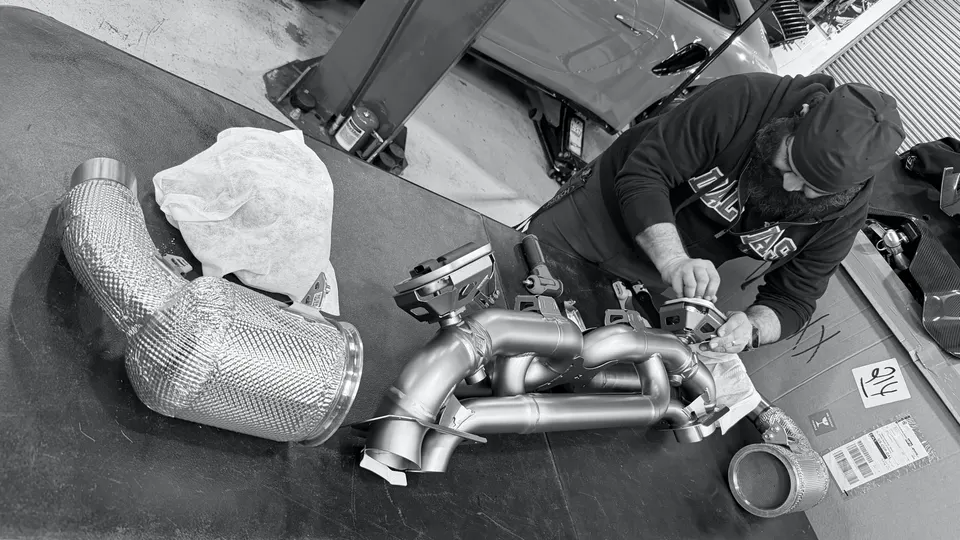 A mechanic working on a performance exhaust system with heat-wrapped components in a car tuning shop.