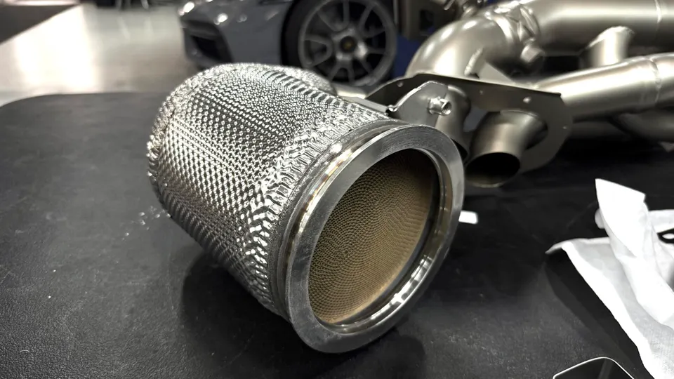 Close-up of a high-flow catalytic converter for a performance exhaust system, with a Lamborghini in the background.