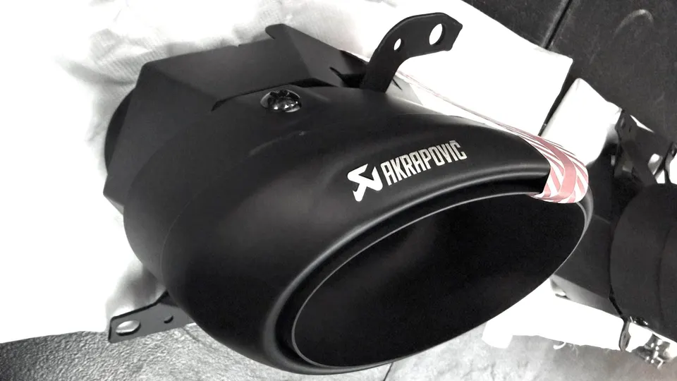 Close-up of a new Akrapovic exhaust tip, matte black finish with white logo, ready for installation.