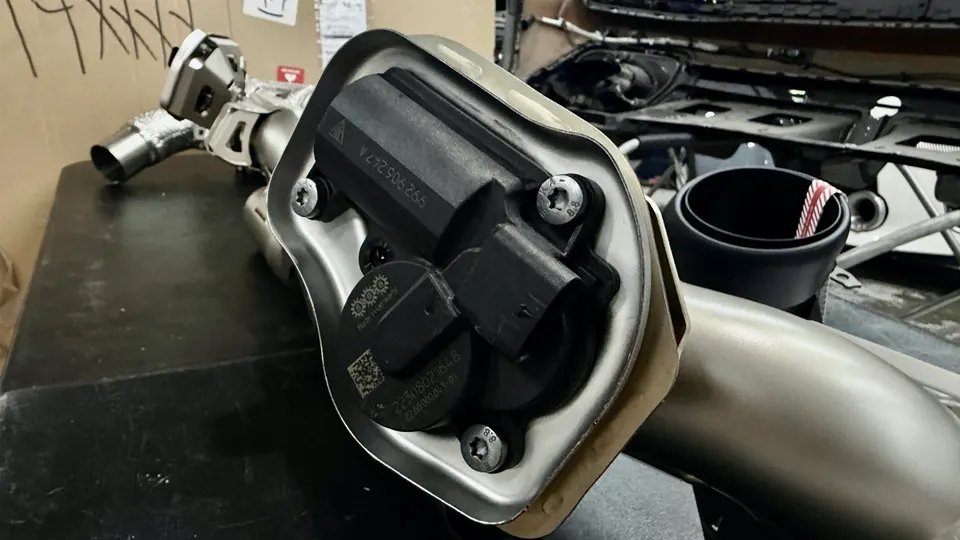 A close-up of a car exhaust system component with an electronic valve on a workbench in a shop.
