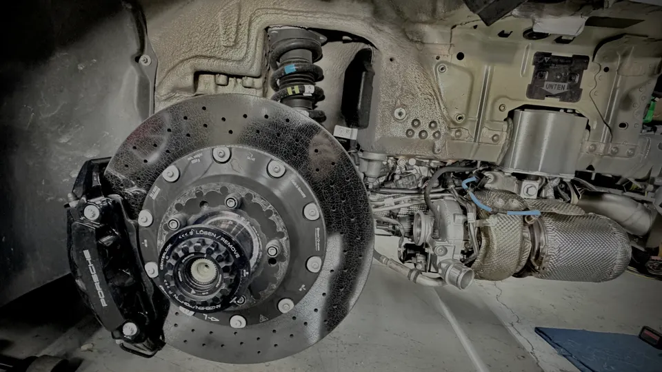 Detailed view of a Porsche's performance brake system, suspension, and turbocharger with heat shield in a shop.