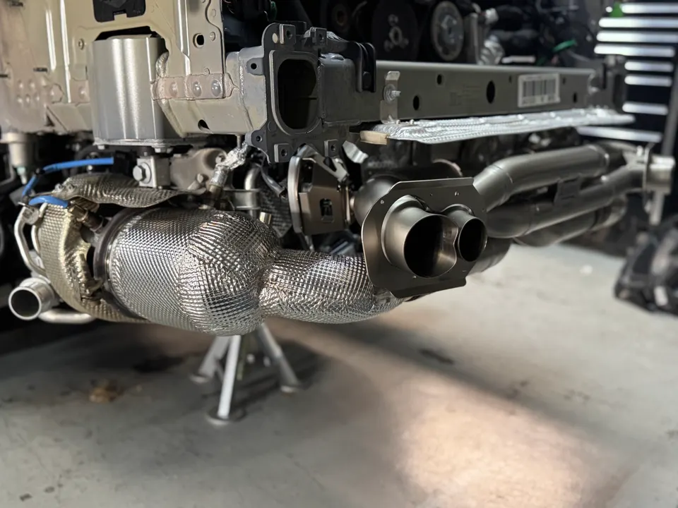 A close-up of a performance exhaust system with heat wrap and dual tips being installed on a car in a shop.
