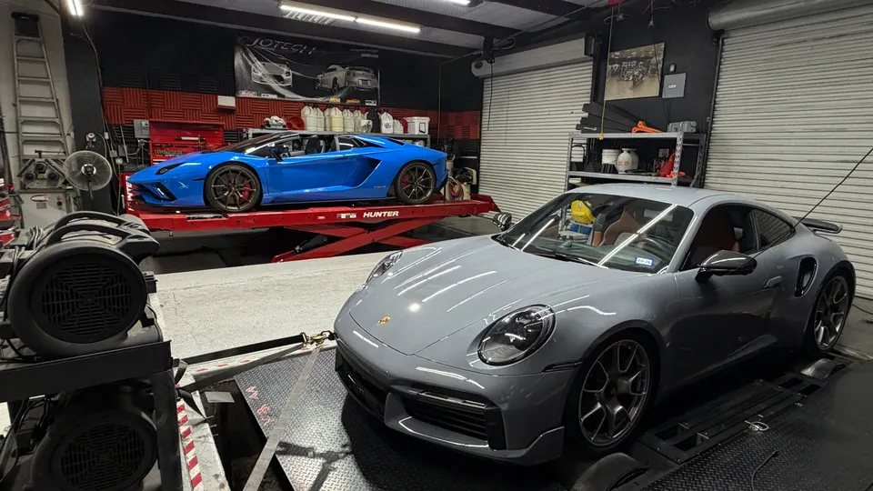 A grey Porsche 911 on a dyno with a blue Lamborghini Aventador on a car lift in a performance tuning shop.