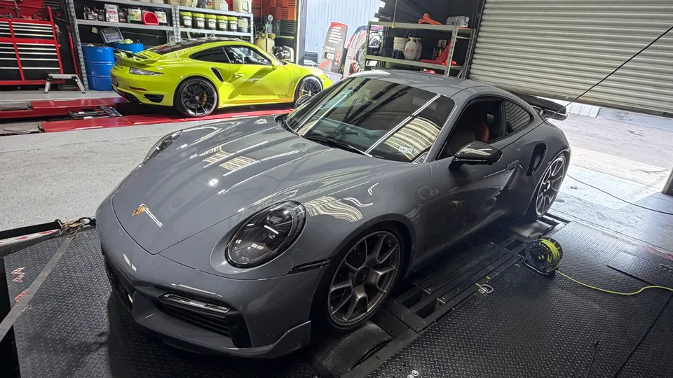 A grey Porsche 911 on a dynamometer with a yellow Porsche 911 in the background at a performance tuning shop.