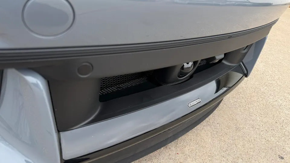 Close-up of a grey car's front bumper featuring a mesh grille, parking sensor, and a Jotech badge.