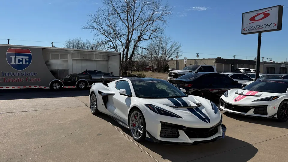 White Corvette Z06 with blue stripes parked outside Jotech Motorsports with other performance cars and a car trailer.