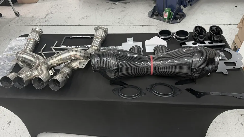 BoostLogic carbon fiber intake and stainless steel exhaust system laid out on a table in a performance shop.