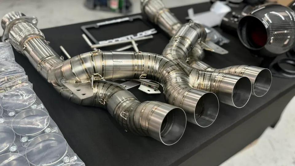 Titanium exhaust downpipes and carbon fiber intake system laid out on a table in a performance shop.