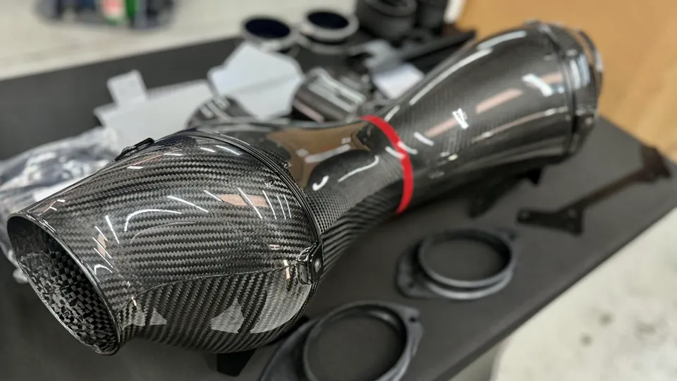Close-up of a carbon fiber aftermarket intake system with a red accent band, displayed on a table in a shop.