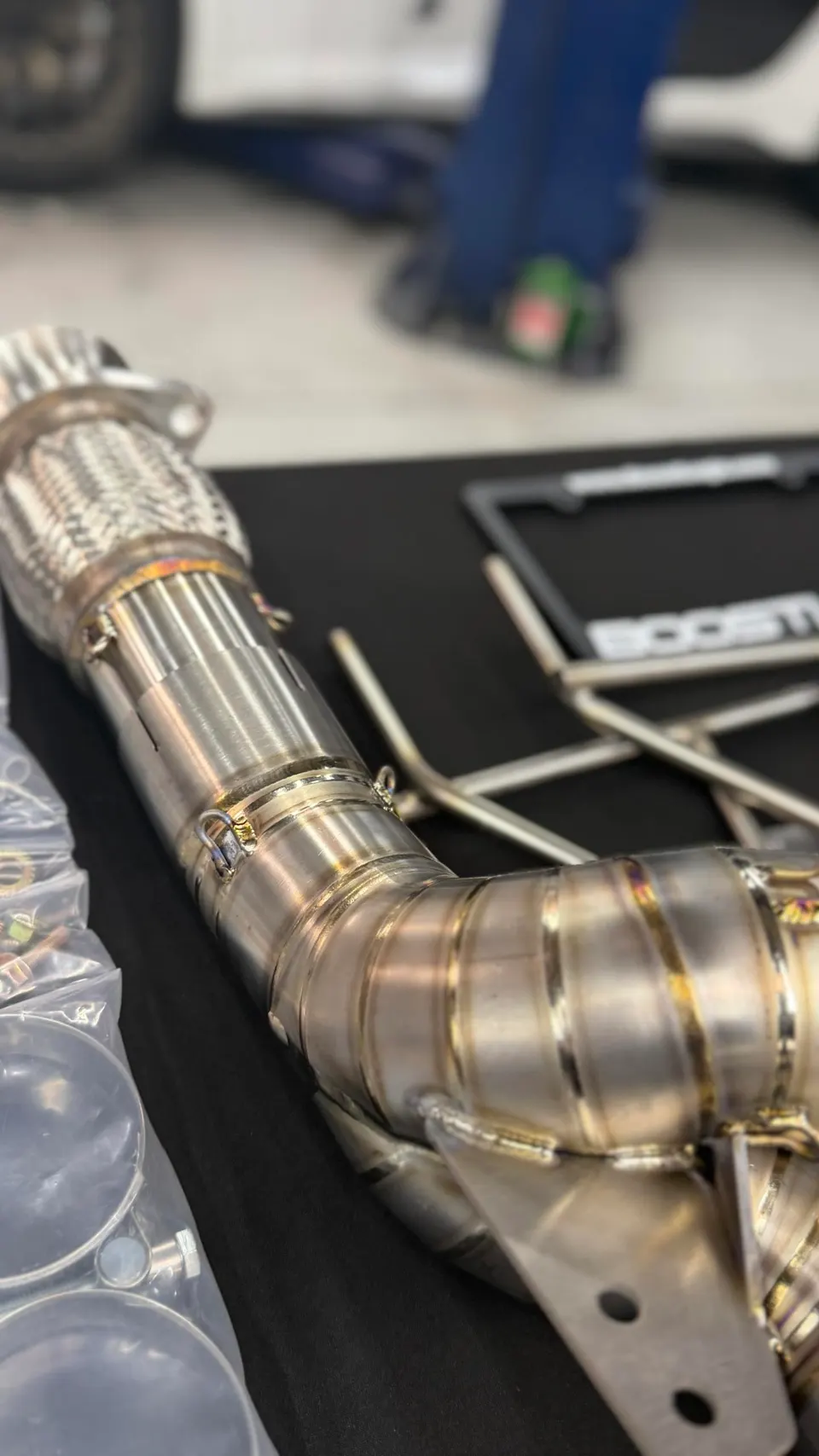 Close-up of a new stainless steel exhaust downpipe with visible welds and a flexible section, ready for installation in a shop.
