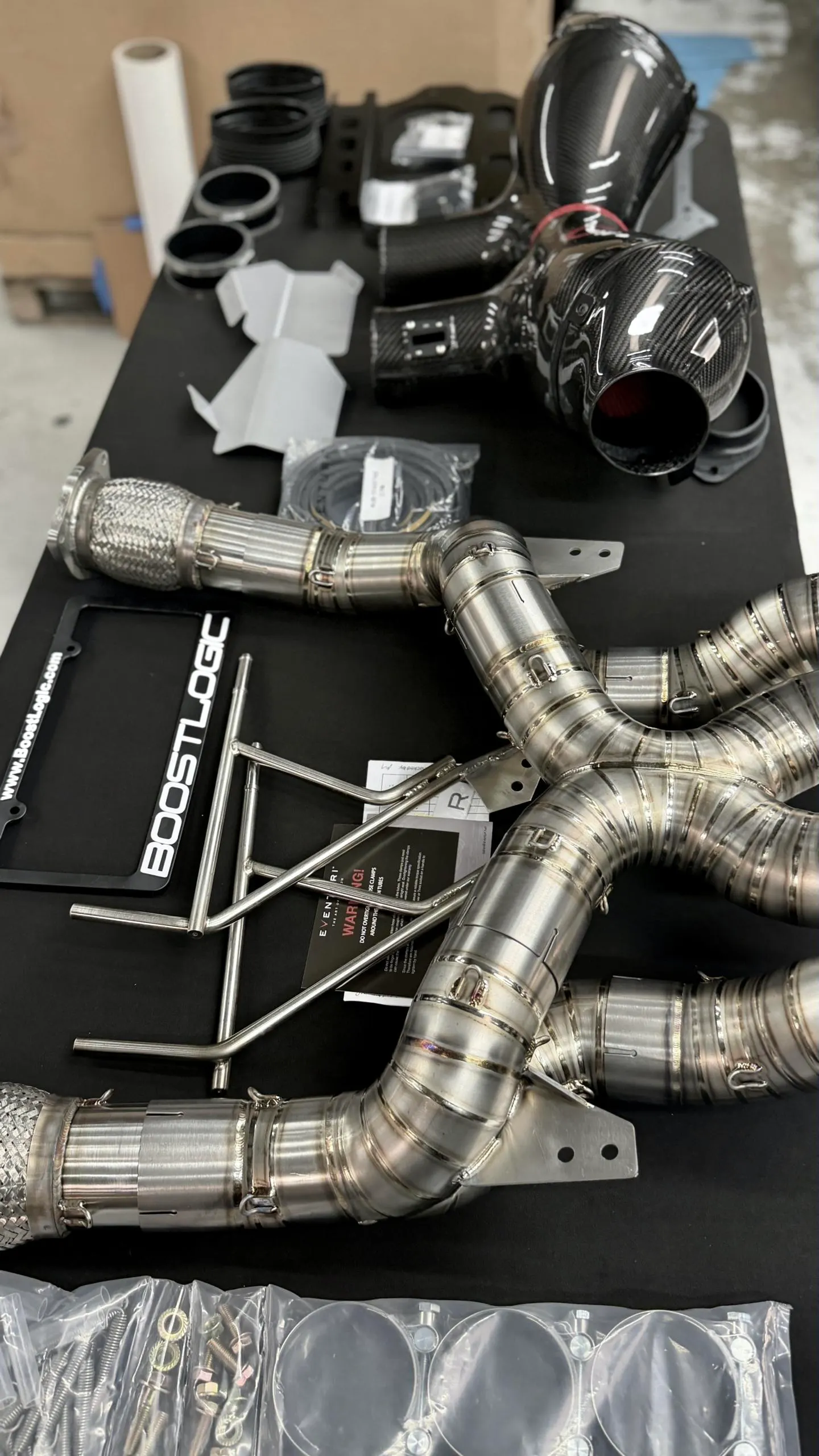 High-performance carbon fiber intake and titanium exhaust system components from BoostLogic laid out on a table.