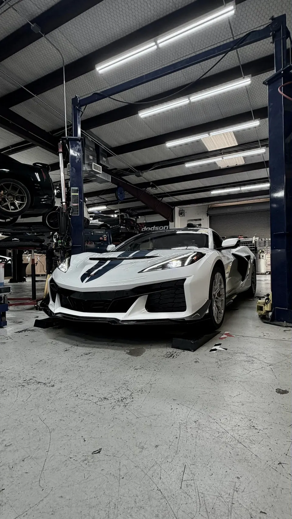 A white Chevrolet Corvette C8 Z06 with blue racing stripes parked in a performance tuning shop with a car on a lift.