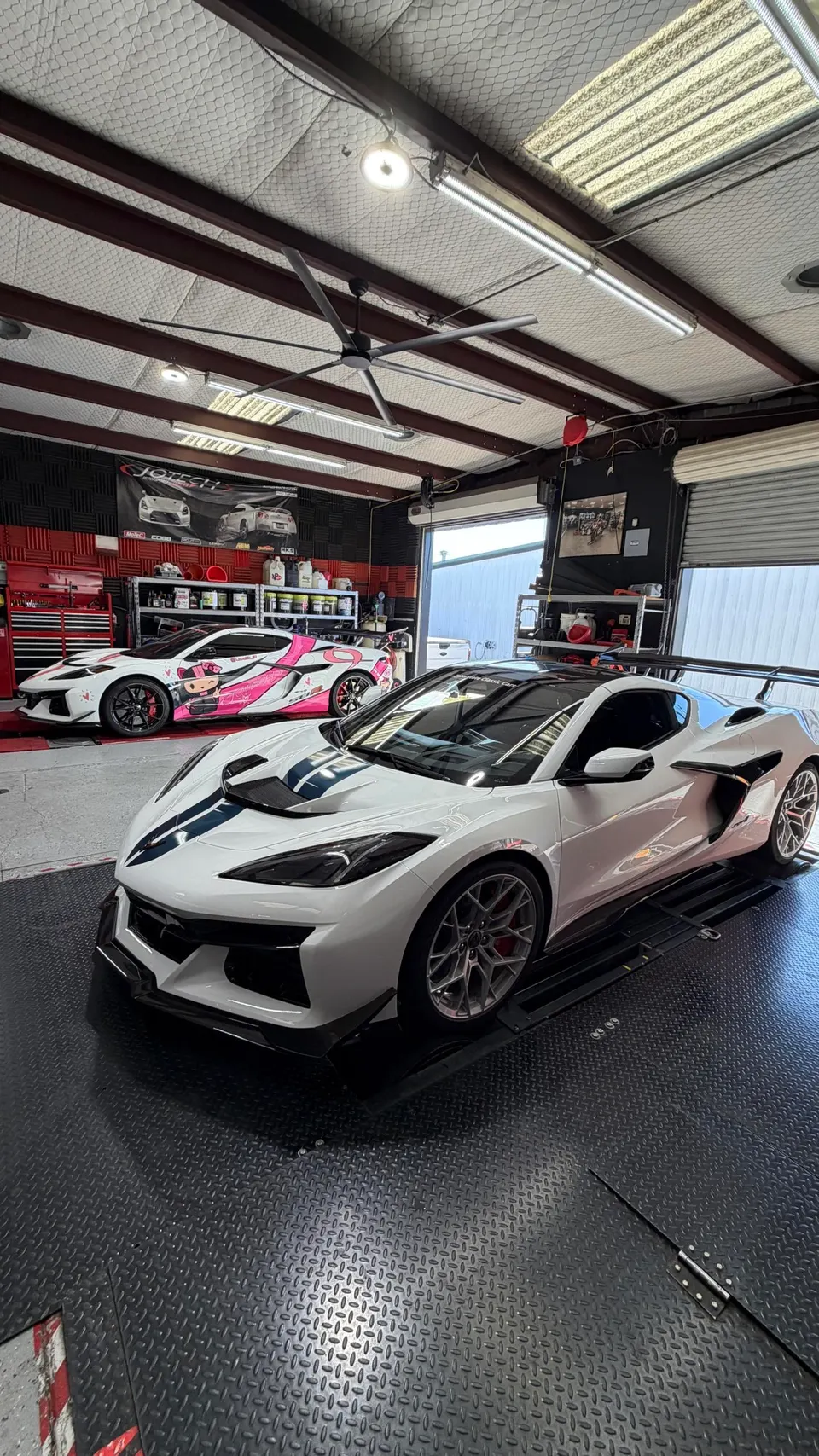 Two modified Chevrolet C8 Corvettes, one white with blue stripes and another with a pink anime wrap, inside a performance tuning shop with a dyno.