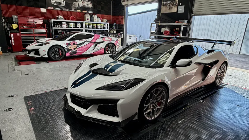 Two custom Chevrolet C8 Z06 Corvettes, one white with blue stripes and a large wing, the other pink wrapped, inside a performance shop with a dyno.