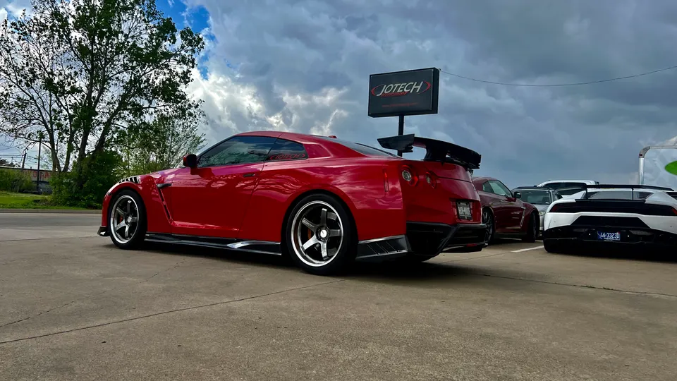 Red Nissan GT-R with aftermarket wheels and a large rear wing parked outside Jotech Motorsports, with a white Lamborghini in the background.