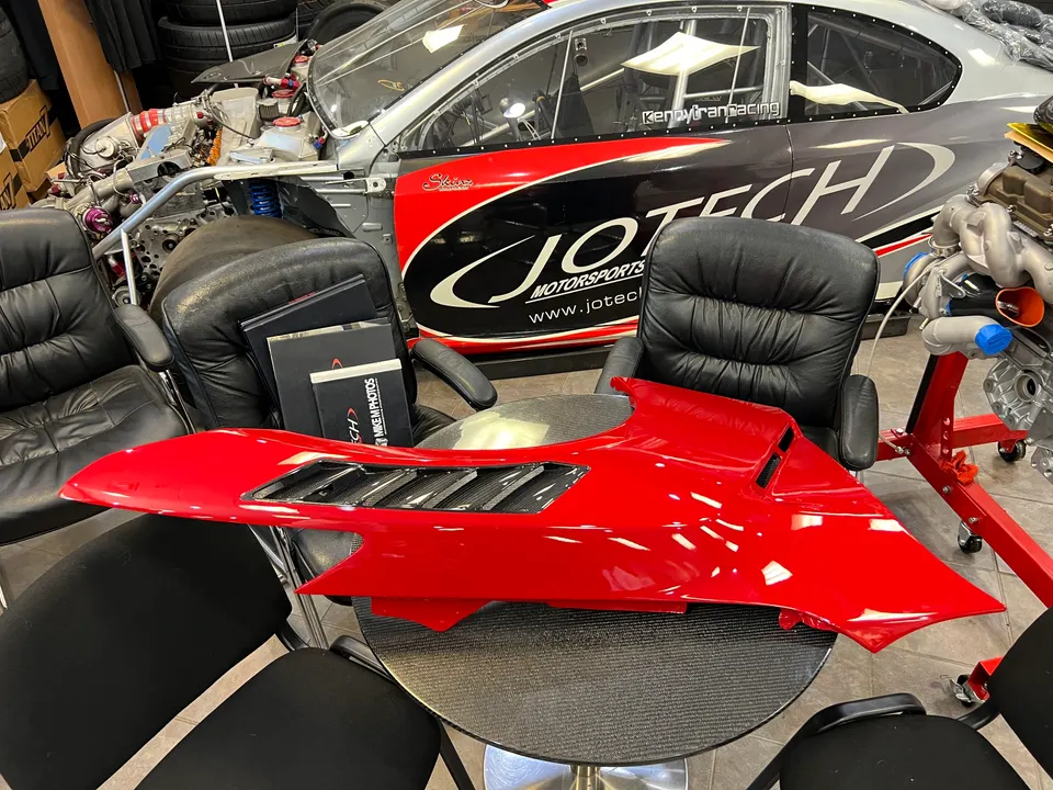 A red vented body panel on a table in the Jotech Motorsports shop with a Nissan GT-R race car chassis in the background.