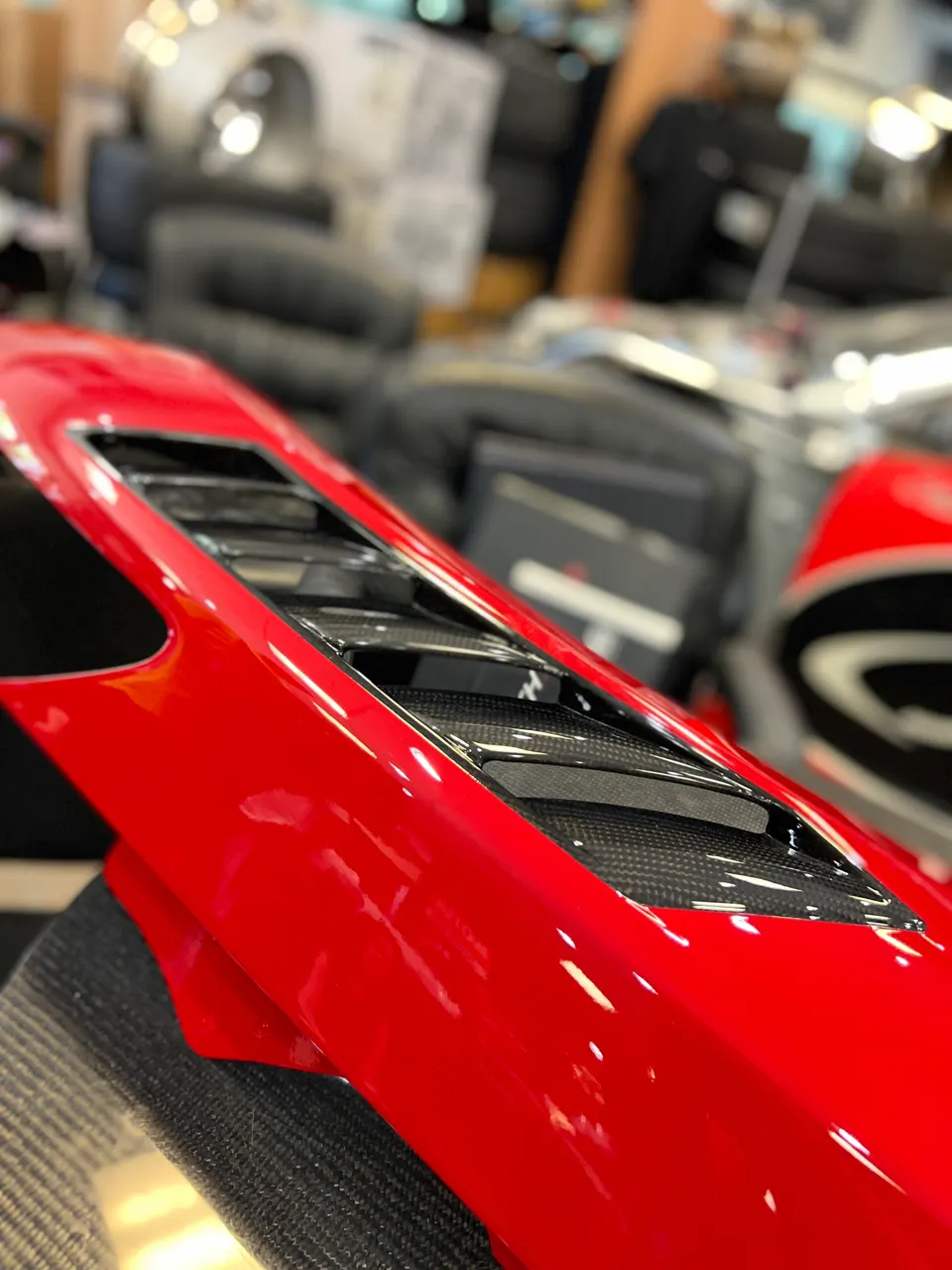 A close-up of a glossy red car body panel featuring carbon fiber vents, likely a hood or fender component, in a shop setting.