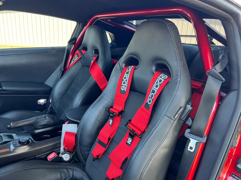 Interior view of a Nissan GT-R with black racing seats, red Sparco harnesses, and a red roll cage.