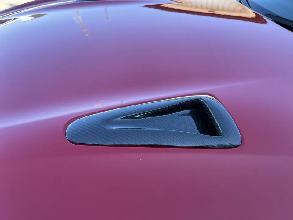 Close-up of a red car's hood featuring a carbon fiber vent for enhanced aerodynamics.