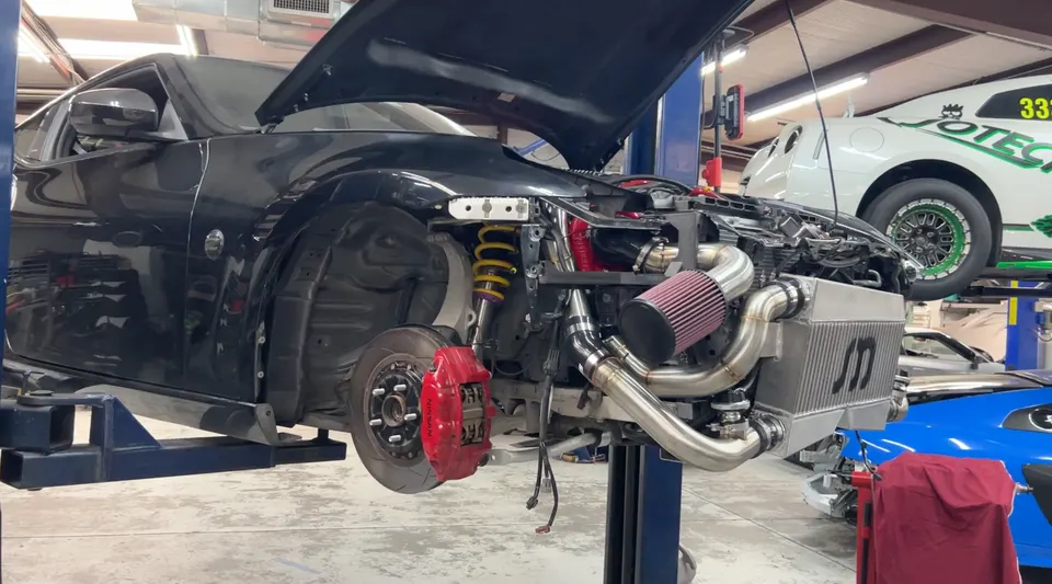 Black Nissan 370Z on a lift in a shop, undergoing a turbo kit installation with a large intercooler and aftermarket suspension.