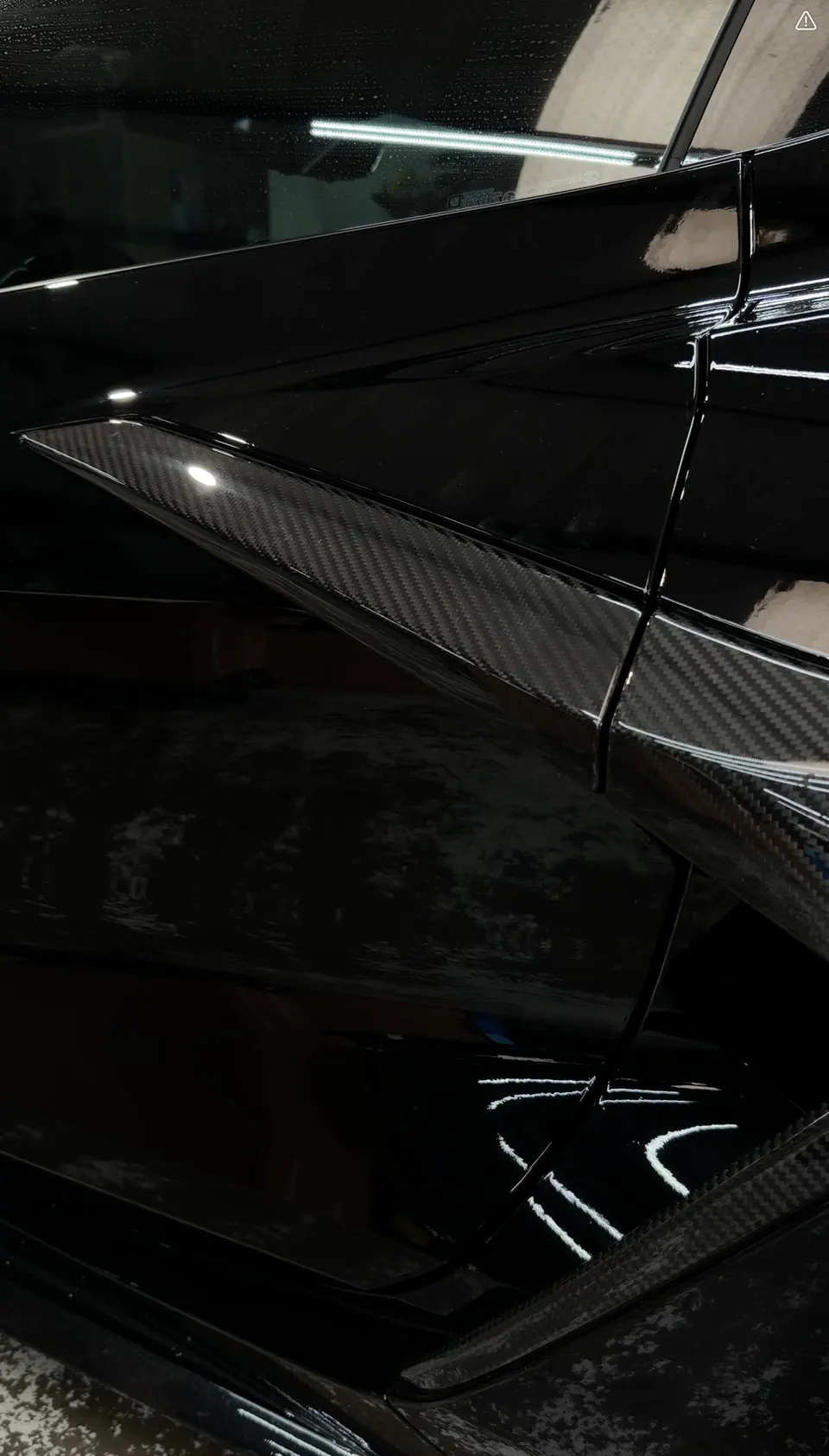 A close-up of a black car's carbon fiber side skirt, showcasing the weave pattern and glossy finish.