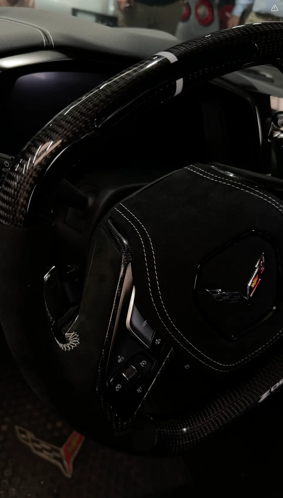A close-up of a custom carbon fiber and Alcantara steering wheel with white stitching and a Corvette logo.
