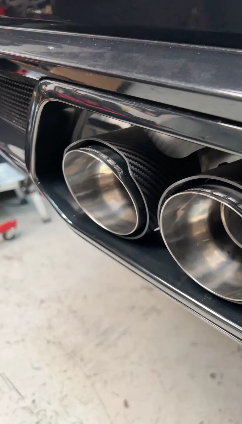 Close-up of aftermarket carbon fiber exhaust tips on a performance car, likely in a shop setting.