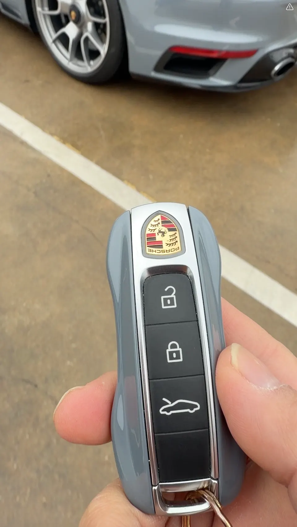 A person holding a grey Porsche car key fob with a grey Porsche car in the background, showing the wheel and rear bumper.
